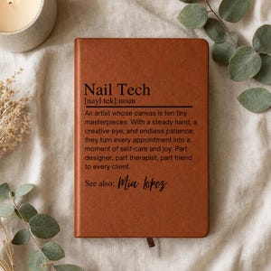 Könnte beinhalten: Ein braunes, ledergebundenes Notizbuch mit der schwarz aufgedruckten Definition von Nail Tech, die einen Künstler mit kreativem Auge und ruhiger Hand beschreibt, gefolgt vom Text See also: Mia Lopez in Schreibschrift.