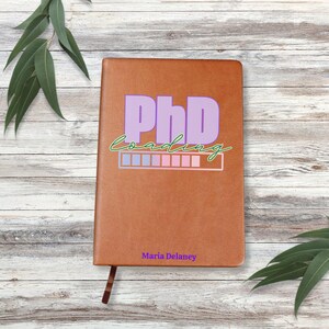 May include: A brown leather journal with the text "PhD Loading" in purple and green lettering. The text is accompanied by a progress bar with alternating pink and blue squares. The journal is personalized with the name "Maria Delaney".