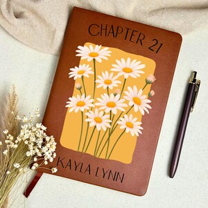 May include: A brown leather journal with the text "CHAPTER 21" and "KAYLA LYNN" printed on the cover. The journal features a floral design of white daisies with yellow centers on a yellow background. A pen rests beside the journal.