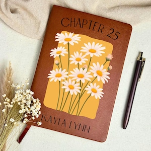 May include: A brown leather-bound journal with the text "CHAPTER 25" and "KAYLA LYNN" printed on the cover. The journal features a floral design of white daisies with yellow centers on a yellow background. A dark pen rests beside the journal.