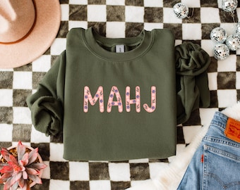 May include: An olive green sweatshirt with the word "MAHJ" in a floral design. The letters are pink with blue and white flower accents. The sweatshirt is on a checkered surface with a hat, plant, and jeans.