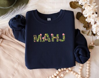 May include: Navy blue sweatshirt with the word "MAHJ" in a floral design. The letters are filled with a pattern of green leaves and pink flowers. The sweatshirt is displayed on a cream-colored surface with decorative elements.