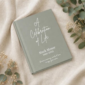 May include: A sage green hardcover book with white text that reads A Celebration of Life, Mark Moser, 1949-2025, and Share a memory, words of love or sign your name. The book has a smooth, matte finish and is positioned at a slight angle.