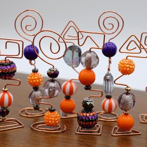 Puede incluir: Un conjunto de 10 esculturas de alambre con temática de Halloween decoradas con cuentas naranjas, moradas y blancas. Cada escultura presenta una forma geométrica diferente, incluyendo un triángulo, un cuadrado y un círculo.