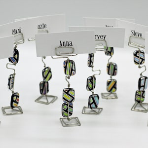 Puede incluir: Un conjunto de ocho porta tarjetas de lugar hechos de alambre plateado con cuentas de vidrio de colores. Las tarjetas de lugar tienen nombres escritos en ellas, incluyendo Roxanne, Mark, Anna, Steve, Isaac, y otros.