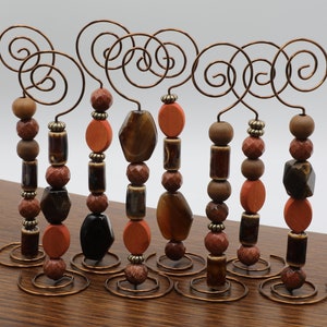 Puede incluir: Conjunto de ocho portafotos de alambre de cobre con cuentas marrones, naranjas y negras. Cada portafotos tiene una parte superior en espiral y una base con un pequeño círculo de alambre de cobre.