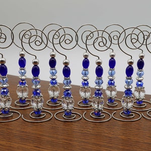 Puede incluir: Doce porta tarjetas de mesa de alambre plateado con cuentas de vidrio azul y transparente. Cada porta tarjetas tiene una base en espiral y un gancho en la parte superior.