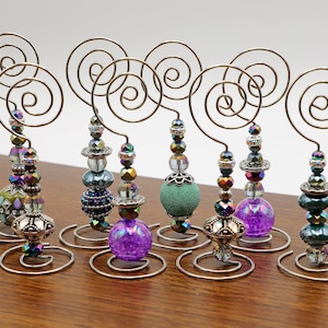Puede incluir: Ocho porta tarjetas de mesa de alambre decorativos con cuentas iridiscentes y una variedad de acentos de vidrio y metal de colores. Los soportes están hechos de alambre y tienen un diseño en espiral.