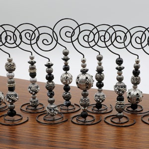 Puede incluir: Un conjunto de diez sujetamemorias de alambre negro con detalles de cuentas decorativas. Cada soporte presenta una parte superior en espiral y una base circular. Las cuentas son plateadas, negras y grises, con diversas texturas y patrones.