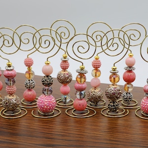 Puede incluir: Conjunto de 10 portafotos de alambre dorado con cuentas rosas y doradas. Cada portafotos tiene una parte superior de alambre en espiral y una base con una variedad de cuentas rosas y doradas.