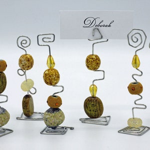 Puede incluir: Seis porta tarjetas de alambre plateado con diferentes diseños. Cada porta tarjetas está decorado con cuentas marrones, amarillas y blancas. Un porta tarjetas tiene una tarjeta con el nombre "Deborah" escrito en ella.
