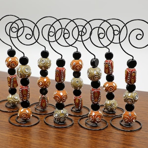 Puede incluir: Diez soportes de exhibición de metal negro con un diseño en espiral, cada uno con un collar de cuentas con cuentas de color naranja, marrón y blanco.