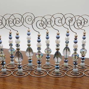 Puede incluir: Doce soportes de alambre plateado con cuentas de vidrio azul e iridiscente. Los soportes están decorados con un diseño en espiral en la parte superior.