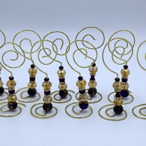 Puede incluir: Una colección de colgadores decorativos dorados. Cada colgador presenta una parte superior en espiral y un tallo con cuentas ámbar y moradas oscuras. La base de cada colgador es una pequeña espiral circular.