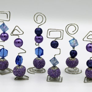 Puede incluir: Seis portafotos de alambre plateado con cuentas moradas y azules. Cada portafotos tiene una forma diferente en la parte superior, incluyendo un cuadrado, un círculo y un triángulo.