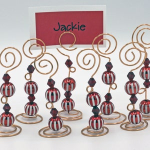 Puede incluir: Conjunto de ocho porta tarjetas de mesa de alambre dorado con cuentas de rayas rojas y blancas y cuentas de cristal rojo. El porta tarjetas está diseñado para sostener una tarjeta de mesa con el nombre "Jackie" escrito en ella.