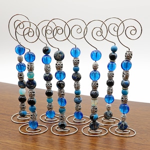 Puede incluir: Diez soportes para adornos de cuentas azules y plateadas. Cada soporte está hecho de una espiral de alambre plateada con una cuerda de cuentas unida. Las cuentas son azules y plateadas y están dispuestas en un patrón aleatorio.