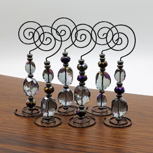 Puede incluir: Seis soportes de adorno de metal negro con cuentas de vidrio iridiscente. Cada soporte tiene un diseño en espiral en la parte inferior.