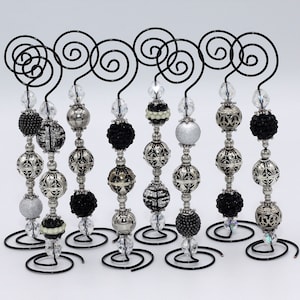 Puede incluir: Conjunto de ocho soportes para adornos de cuentas negras y plateadas. Cada soporte tiene un soporte en espiral de metal plateado y una hebra de cuentas negras, plateadas y blancas.