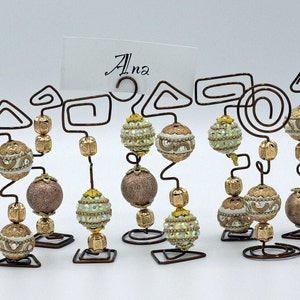 Puede incluir: Conjunto de 10 porta tarjetas de mesa de alambre con cuentas doradas y blancas. Cada porta tarjetas presenta un diseño de alambre único y una cuenta decorativa. La palabra "Ana" está escrita en una tarjeta en el fondo.