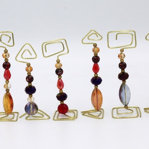 Puede incluir: Ocho soportes de alambre dorado con diferentes formas geométricas, cada uno rematado con una cadena de cuentas de vidrio de colores. Las cuentas son en tonos de rojo, naranja, amarillo, azul y morado.