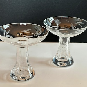 May include: Two clear glass dessert bowls with etched floral designs. The bowls have a wide, shallow top and a stemmed base. The etched designs include flowers and leaves. The bowls are on a white surface with a black background.
