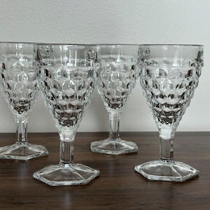 May include: Four clear, vintage wine glasses with a honeycomb pattern. The glasses have a hexagonal base and a long stem. The glasses are made of glass and are displayed on a dark brown wooden surface.
