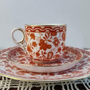 Może przedstawiać: Biały zestaw do herbaty z czerwonym kwiatowym wzorem i złotym obramowaniem. Filiżanka, spodek i talerz mają pasujący wzór. Zestaw stoi na białej powierzchni.