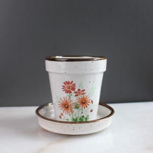 Puede incluir: Maceta de cerámica blanca con platillo a juego. La maceta presenta un diseño floral en tonos naranja y rojo, con hojas verdes. El borde y el borde del platillo tienen un ribete marrón. La maceta está moteada con pequeños puntos marrones.