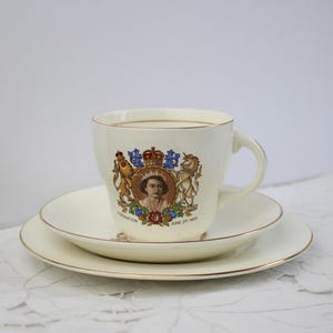 Può includere: Servizio da tè color crema con bordi dorati. La tazza da tè presenta un ritratto e un testo che commemora l'incoronazione del 2 giugno 1953. Il set è esposto su una tovaglia di pizzo bianco.