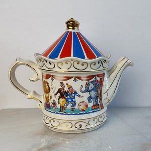 Op de afbeelding: Een keramische theepot met een circusthema en een deksel in de vorm van een rood-blauw gestreepte tent. De body toont een circusdirecteur, een clown, een leeuw en een olifant, afgewerkt met gouden randen op het handvat, de tuit en de voet.