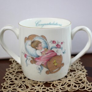 Op de afbeelding: Witte keramische babymok met twee handvatten, met een illustratie van een baby in een wieg. De mok heeft een gouden rand en het woord "Gefeliciteerd" in lichtblauw. Roze rozen en blauwe linten versieren de mok.