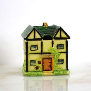 May include: A ceramic cottage-shaped sugar bowl with a green roof, black trim, and a yellow body. The bowl has a small door and windows, and is decorated with green vines and red flowers.
