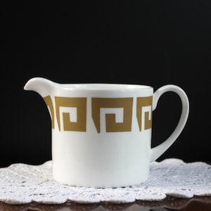 May include: White ceramic creamer with a gold geometric pattern. The creamer has a handle and a spout.