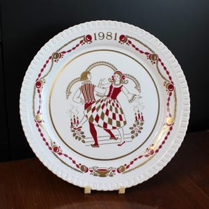 Op de afbeelding: Een wit keramiek kerstbord met een gouden rand en een rood en gouden ontwerp van een dansend stel. Het bord heeft het jaar 1981 erop gedrukt.