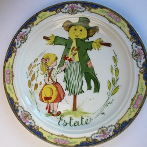 May include: A vintage porcelain plate with a colorful illustration of a girl and a scarecrow. The plate has a yellow, blue, and gold border with floral designs. The text "Estate" is written in cursive on the plate.