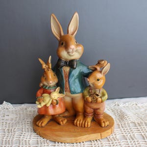 Op de afbeelding: Decoratief beeldje van een konijnenfamilie. De vaderkonijn draagt een groene jas en een vlinderdas, terwijl de kinderen een rode jurk en een overall dragen. Het beeldje staat op een houten voet.