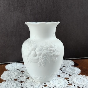 Może przedstawiać: Biały ceramiczny wazon z kwiatowym wzorem reliefowym. Wazon ma szeroki, falisty brzeg i pękaty korpus. Relief przedstawia różne kwiaty i liście. Wazon stoi na białej koronkowej serwetce.