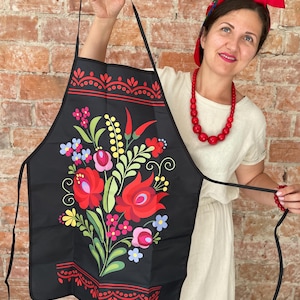 Op de afbeelding: Zwarte schort met een levendig bloemmotief. Het heeft rode, roze en gele bloemen, groene bladeren en blauwe accenten. De schort heeft een decoratieve rand en lange zwarte banden. Achtergrond: bakstenen muur.