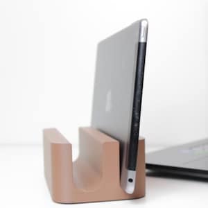 Op de afbeelding: Een bruine tabletstandaard met een tablet. De standaard is gemaakt van een stevig materiaal en heeft een gebogen ontwerp om de tablet rechtop te houden. Een laptop is zichtbaar op de achtergrond.