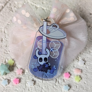 May include: A clear acrylic charm in the shape of a soda can, featuring a blue drink with ice cubes and a cartoon character. The charm is attached to a gold-colored keyring and is set against a pink tulle bow and scattered pastel-colored star and flower-shaped decorations.