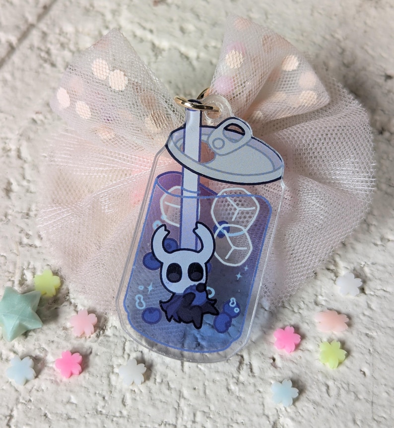May include: A clear acrylic charm in the shape of a soda can, featuring a blue drink with ice cubes and a cartoon character. The charm is attached to a gold-colored keyring and is set against a pink tulle bow and scattered pastel-colored star and flower-shaped decorations.