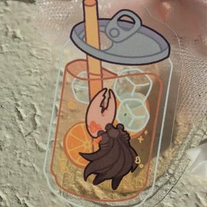 May include: A clear acrylic charm depicting a cartoon drink with ice cubes, an orange slice, and a brown creature. The drink has a straw and a pop-top lid. The charm is rectangular with rounded corners.