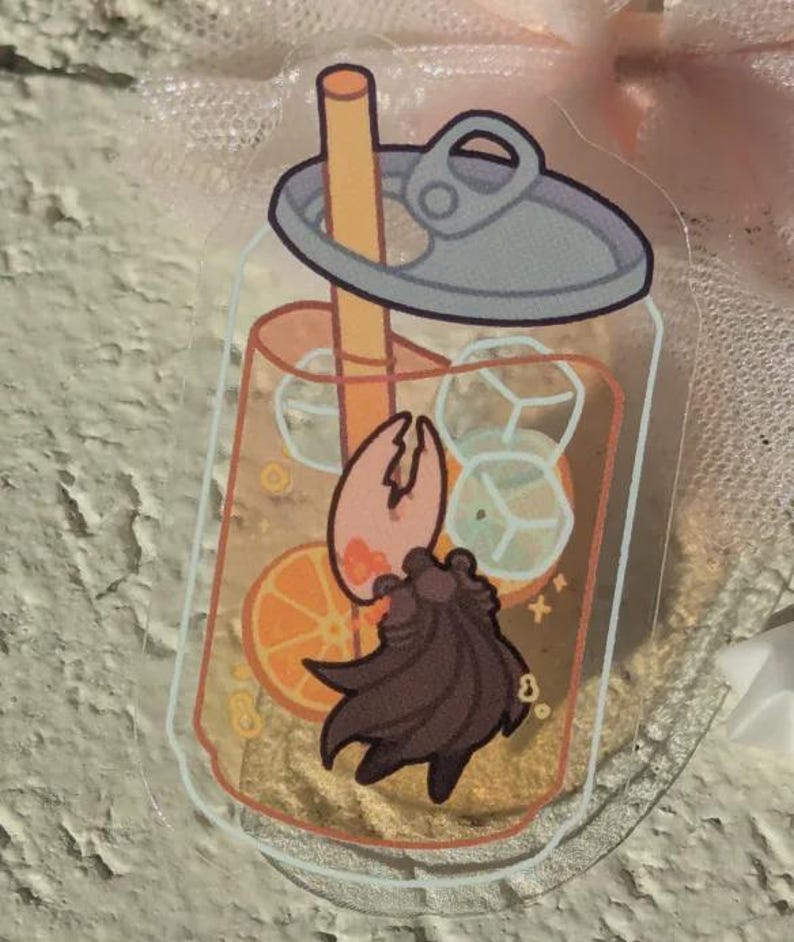 May include: A clear acrylic charm depicting a cartoon drink with ice cubes, an orange slice, and a brown creature. The drink has a straw and a pop-top lid. The charm is rectangular with rounded corners.