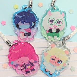 May include: Four character-themed acrylic charms with glitter accents and attached lanyards. Each charm features a different character design with vibrant colors, including blue, pink, green, and yellow. The charms are on a light blue background with star-shaped decorations.