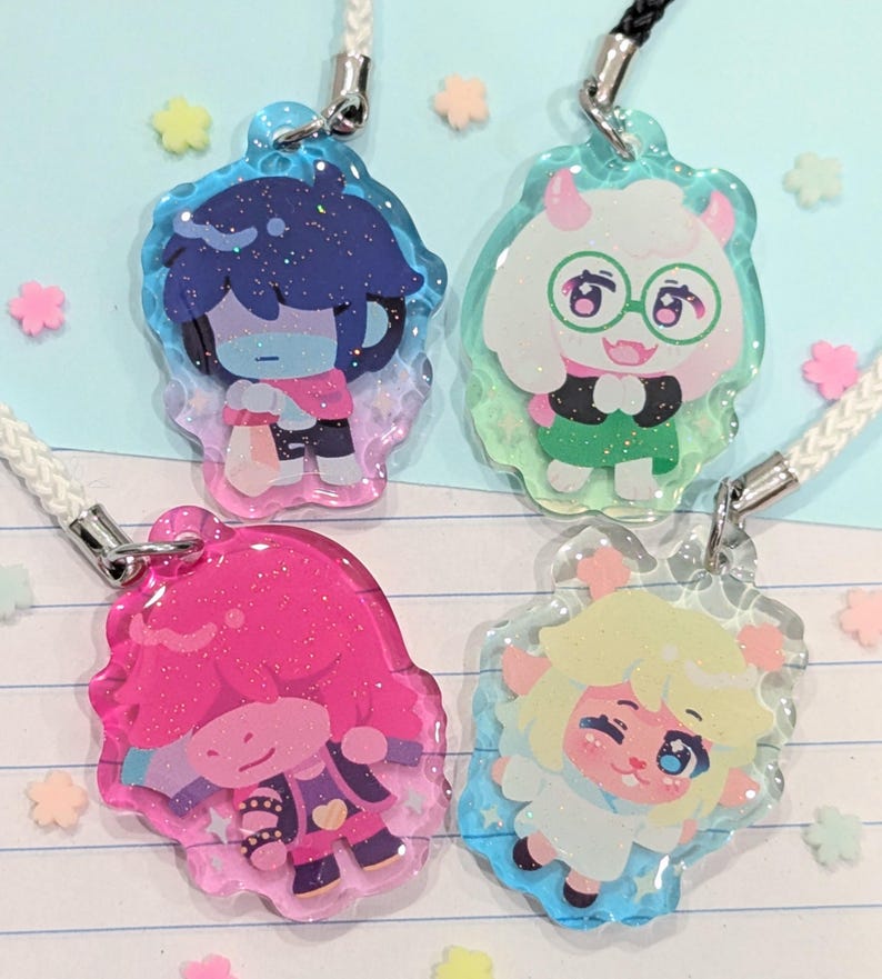 May include: Four character-themed acrylic charms with glitter accents and attached lanyards. Each charm features a different character design with vibrant colors, including blue, pink, green, and yellow. The charms are on a light blue background with star-shaped decorations.