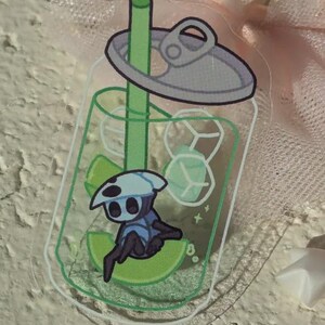 May include: A clear acrylic sticker with a cartoon character inside a soda can design. The character is blue and white, sitting on a lime slice. The can includes ice cubes and a green straw.