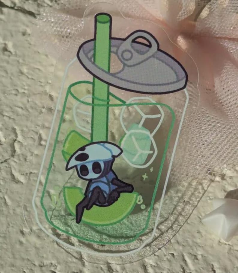 May include: A clear acrylic sticker with a cartoon character inside a soda can design. The character is blue and white, sitting on a lime slice. The can includes ice cubes and a green straw.