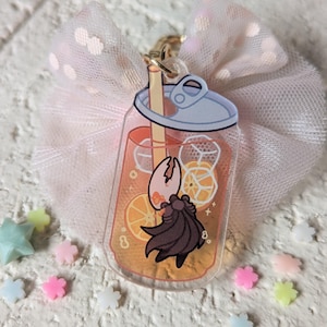 May include: A clear acrylic charm in the shape of a soda can, filled with orange liquid, ice cubes, and orange slices. A cartoon crab is inside the can. The charm is attached to a gold-colored keychain and a pink bow.