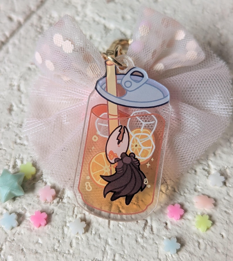 May include: A clear acrylic charm in the shape of a soda can, filled with orange liquid, ice cubes, and orange slices. A cartoon crab is inside the can. The charm is attached to a gold-colored keychain and a pink bow.